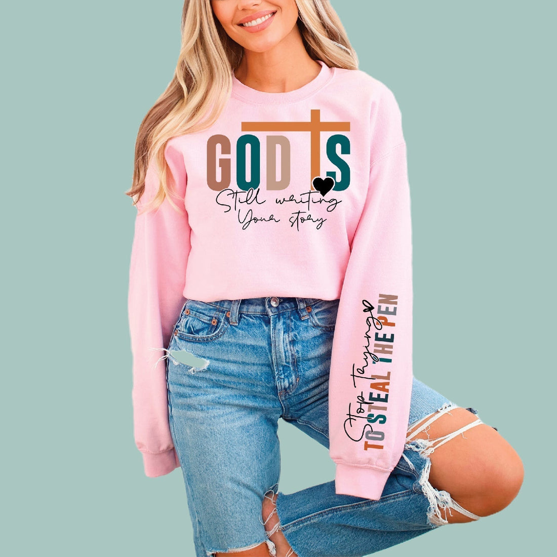 Woman wearing pink Christian sweatshirt with 'God Is Still Writing Your Story' and cross graphic, sleeve reads 'Stop Trying to Steal the Pen' — faith apparel