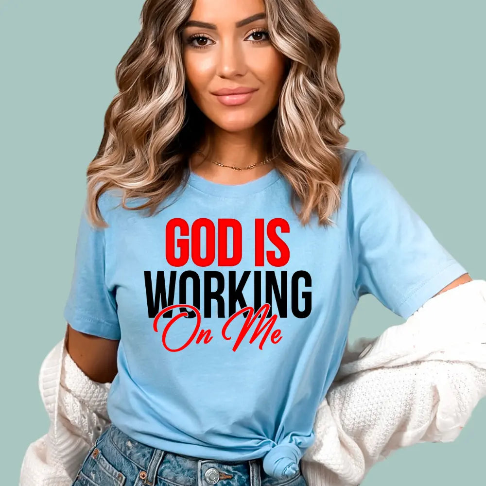 Light blue Christian graphic tee featuring 'God Is Working On Me' in modern bold and script typography.