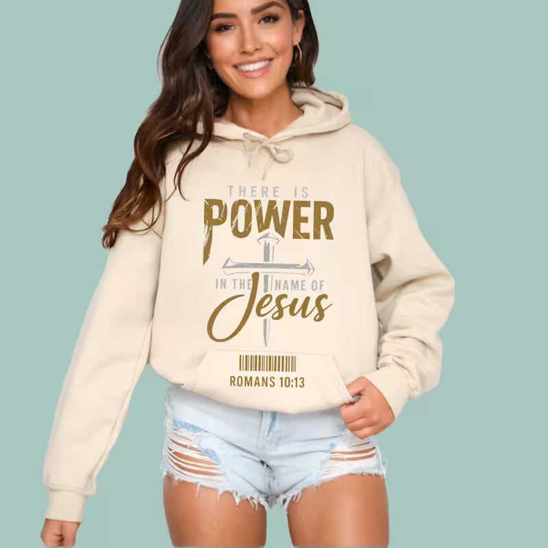 Woman wearing a tan "There Is Power in The Name of Jesus" hoodie with gold and silver graphic.