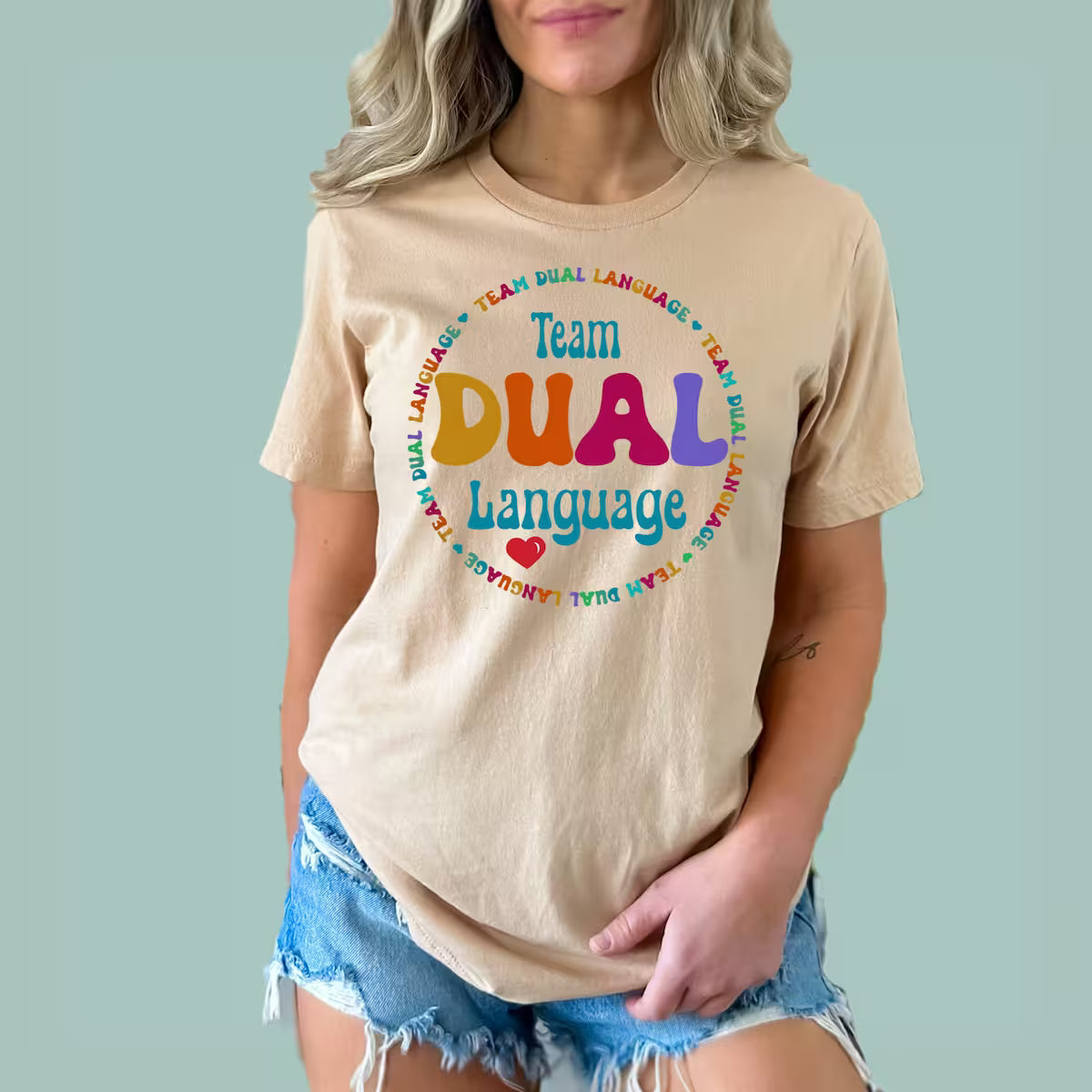 Woman wearing a sand-colored Team Dual Language Teacher tee featuring a colorful bilingual education graphic.