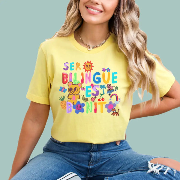 Person wearing a yellow t-shirt with colorful text ser billingue es bonito and graphics on a light blue background